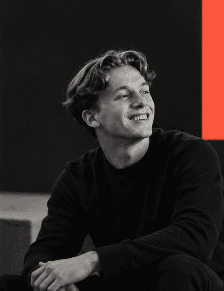 Portrait en noir et blanc de Maxime Derbigny, souriant, devant un fond sombre ponctué d'un aplat vermillon.