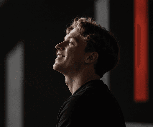 Maxime Derbigny de profil, regard tourné vers la lumière, sur fond sombre.